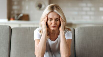 middle aged blonde woman sits on couch at living room holding her head with her hands, feels unhappy because of headache, personal troubles, illness or bad news, she need psychological or medical support
