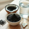 charbon végétal : poudre noire dans un bol en céramique à côté de coques de noix de coco et d'un verre d'eau